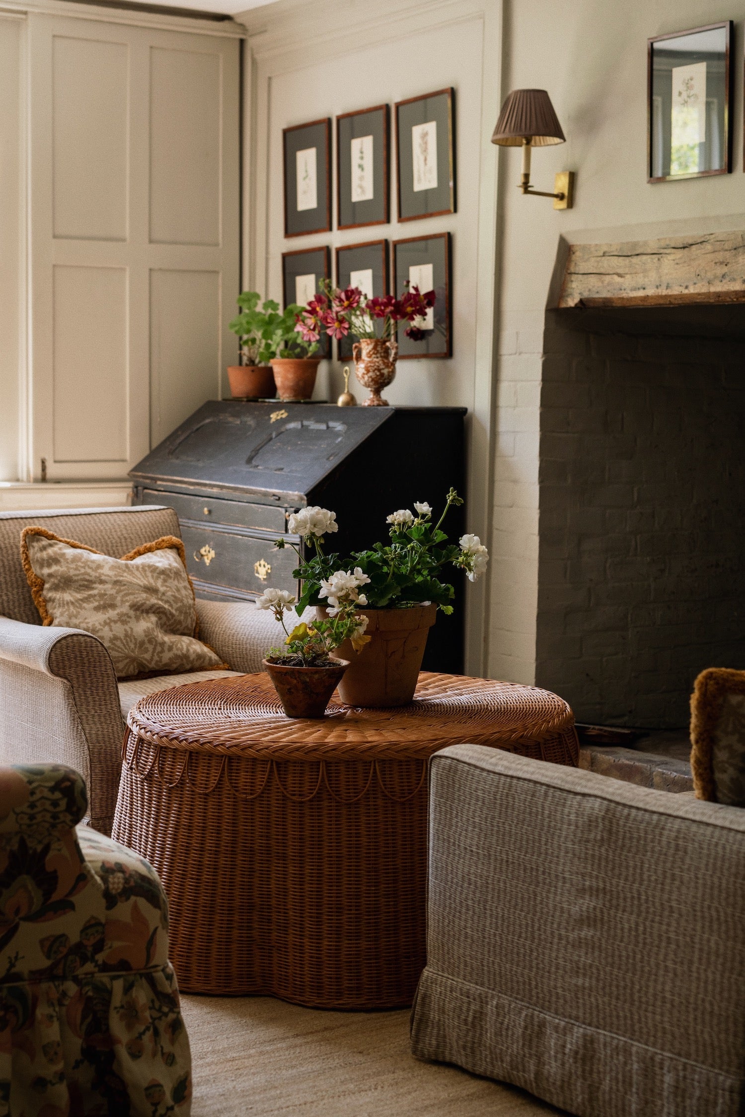 A cozy living room with cushioned chairs, framed art, a fireplace with a rustic mantel, and the Adeline Coffee Table by Sharland England holding potted flowers.