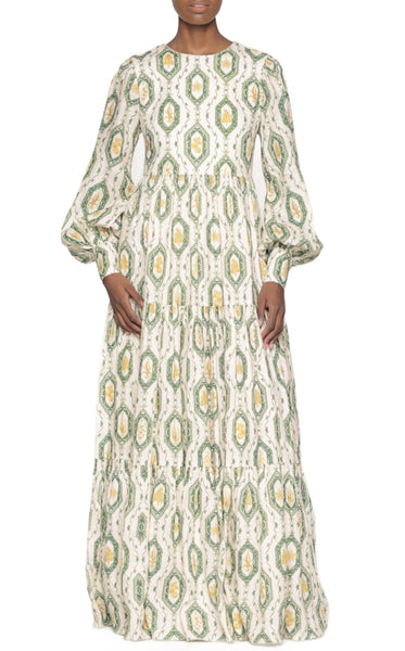 A person wearing an Agua by Aguabendita Nuez Maxi Dress, a long-sleeved, high-necked, flowing linen dress with a green and white patterned design, posing against a plain background.