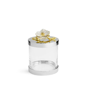 The Michael Aram Orchid Canister is a clear cylindrical glass jar from the Orchid Collection, topped with a silver-colored lid featuring a white flower and gold branch for subtle luxury, purity, and grace.
