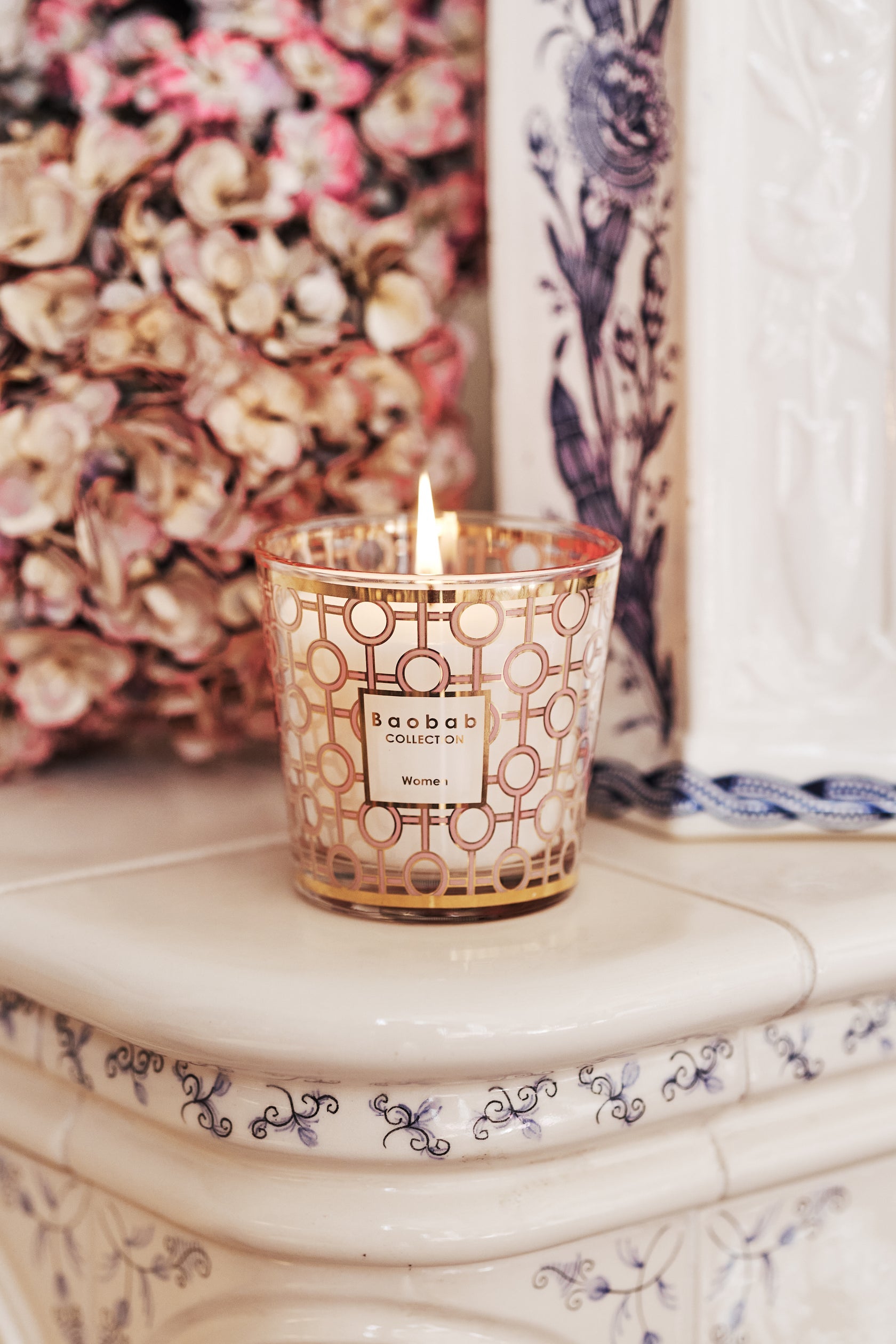 A lit Baobab Women Body Wellness Collection candle by Baobab sits in a decorative glass holder on an ornate white ceramic surface, releasing a floral scent with dried flowers in the background.