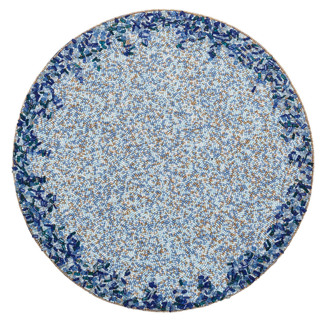 The Kim Seybert Lagoon Placemat showcases a circular mosaic design with a densely patterned blue and brown center accented by cobalt blue beads, bordered by deep ocean-inspired blue and green tones.