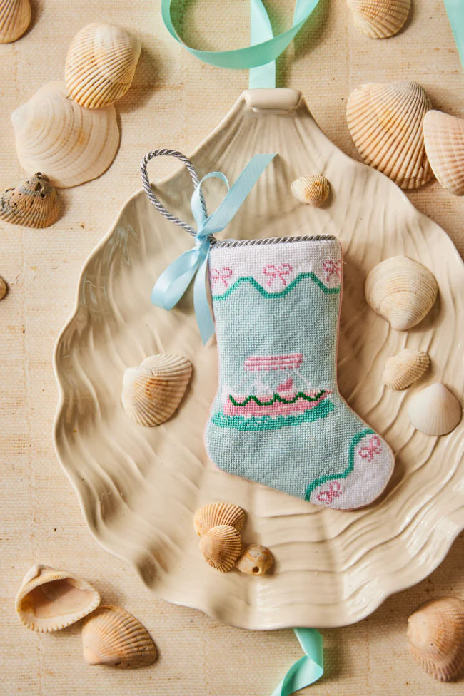 The Palm Yacht Needle Point Stocking by Bauble Stockings, a small pastel nautical Christmas ornament with a blue ribbon, rests on a shell-shaped dish surrounded by assorted seashells.