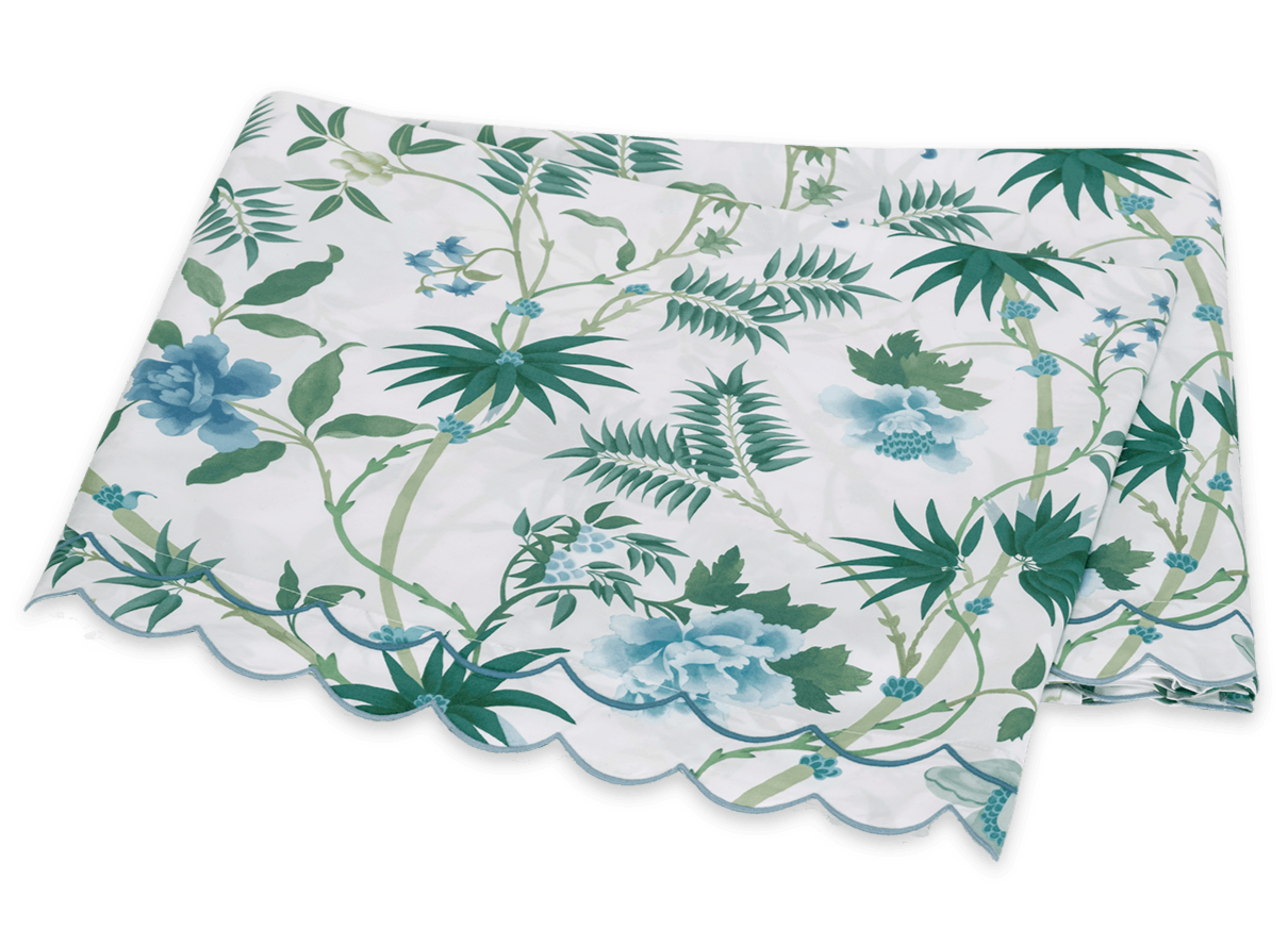 The Matouk Palmyra Bedding Collection, Emerald, features a folded tablecloth with green and blue palm fronds and leafy patterns on a white background, finished with a scalloped edge.