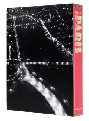 An aerial view of a cityscape at night shows illuminated roads and bridges, while the spine of "Paris by Paris" by Assouline, evoking Belle Époque charm, peeks out.