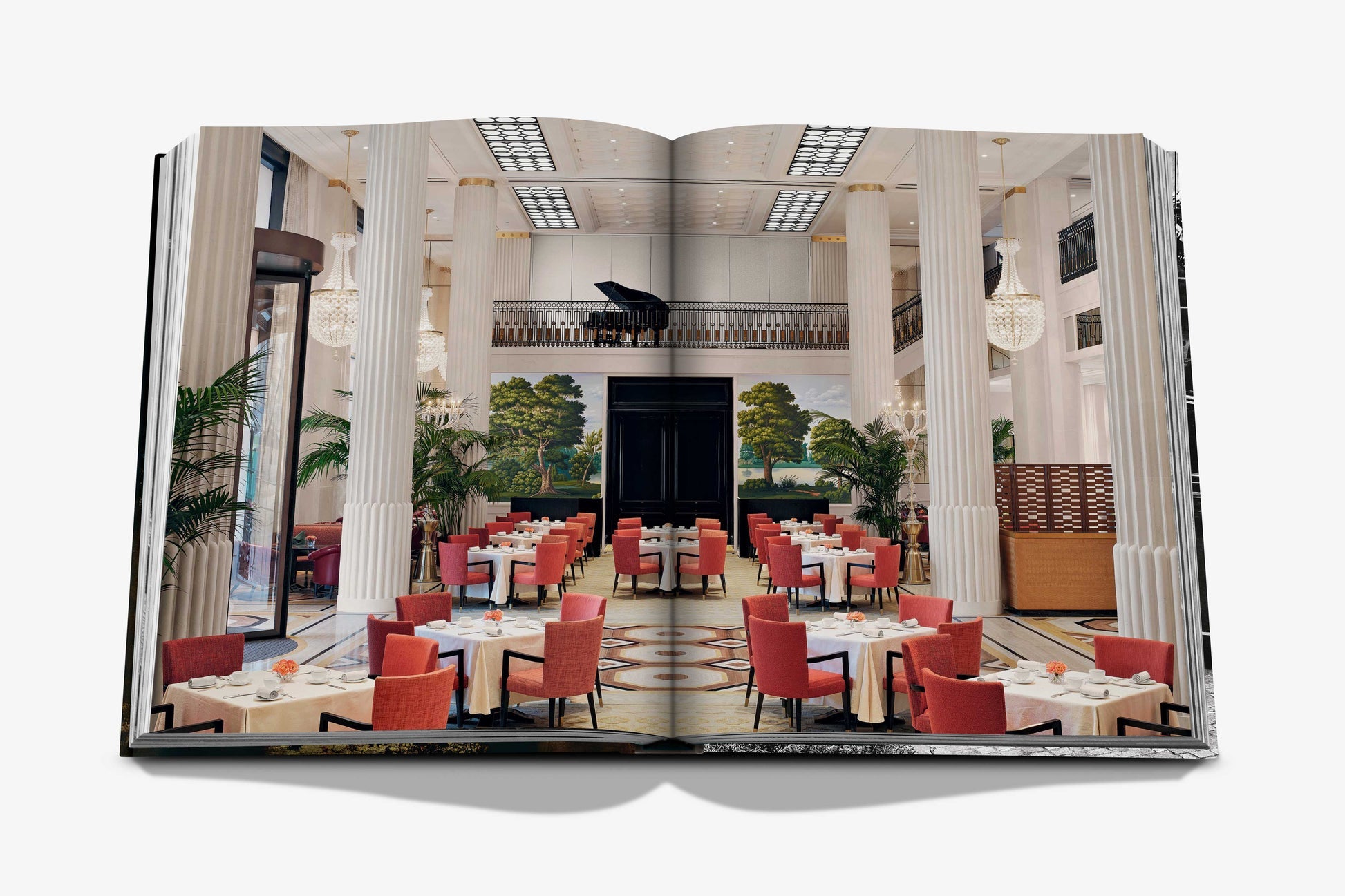 Open book from Assouline featuring The Peninsula London: a Hyde Park hotel with an elegant restaurant, white columns, red chairs, set tables, tall ceilings, and a grand piano on the upper balcony.