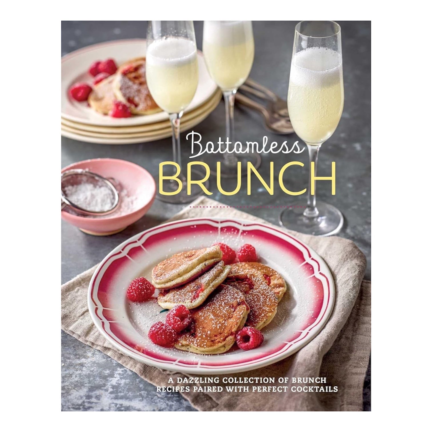A plate of pancakes with raspberries and powdered sugar sits beside two glasses of champagne; the text reads "Bottomless Brunch by Common Ground: A dazzling collection of brunch recipes perfectly paired with cocktails.