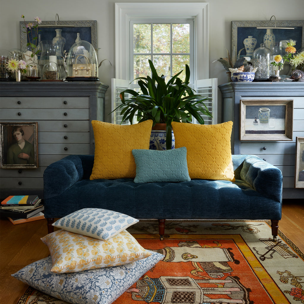 A blue velvet sofa features the John Robshaw Mahin Marigold Decorative Pillow among yellow and blue cushions, set before a window with gray dressers, artwork, and stacked patterned pillows on an ornate rug.