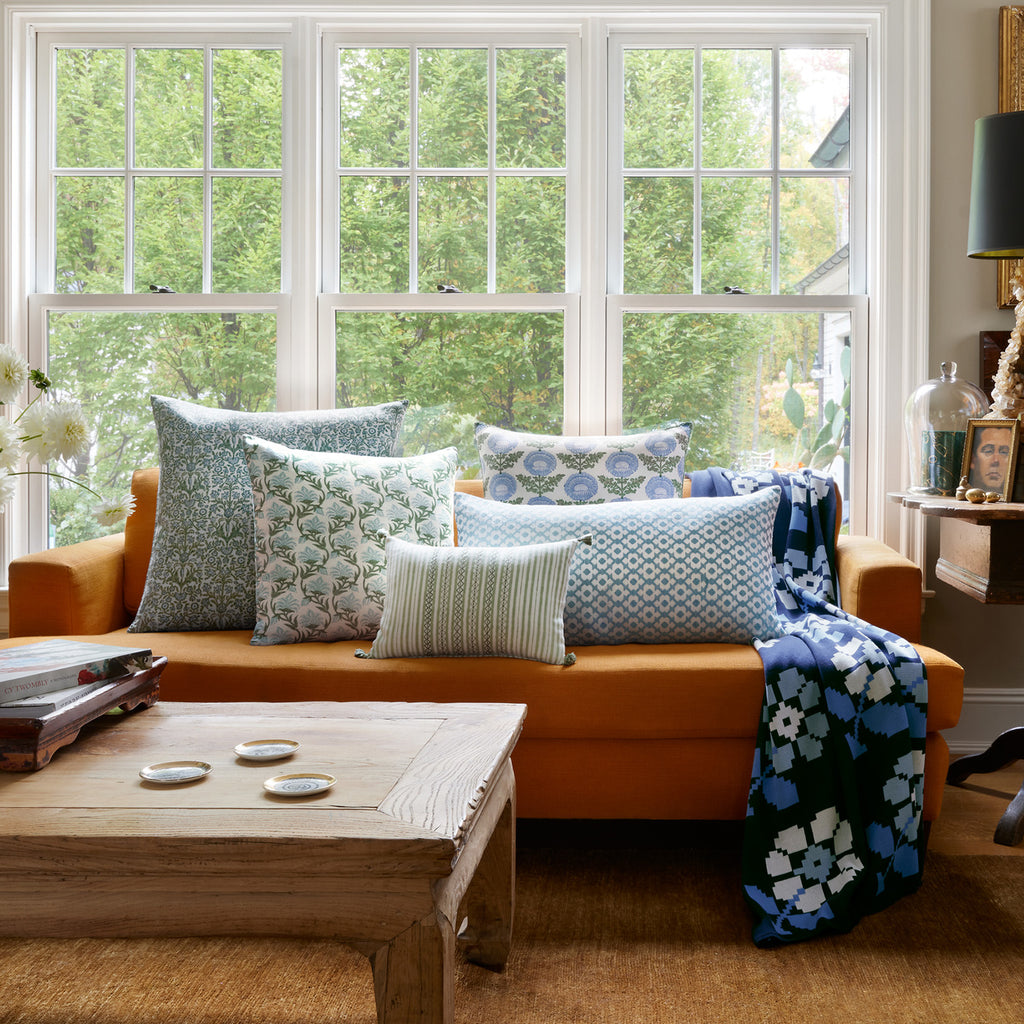 An orange sofa with John Robshaw's Bhuv Sage Kidney Pillows adds a pop of blue and white pattern, some with embroidered stripes, in front of large windows and a wooden coffee table.