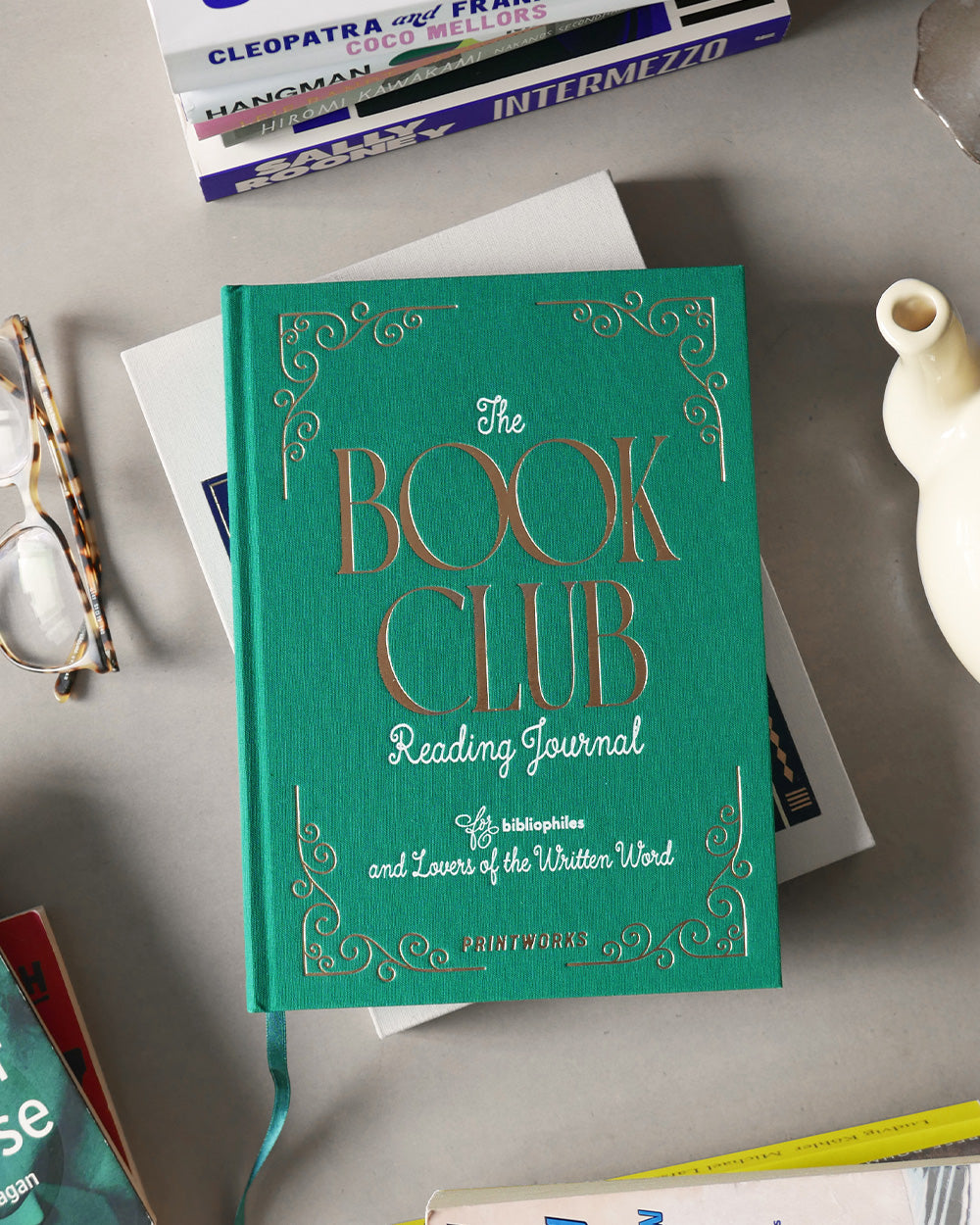 The PrintWorks Book Club Reading Journal, a green hardcover book, rests on a table with glasses, a teapot, and other books—ideal for tracking your reading progress and writing book reviews.