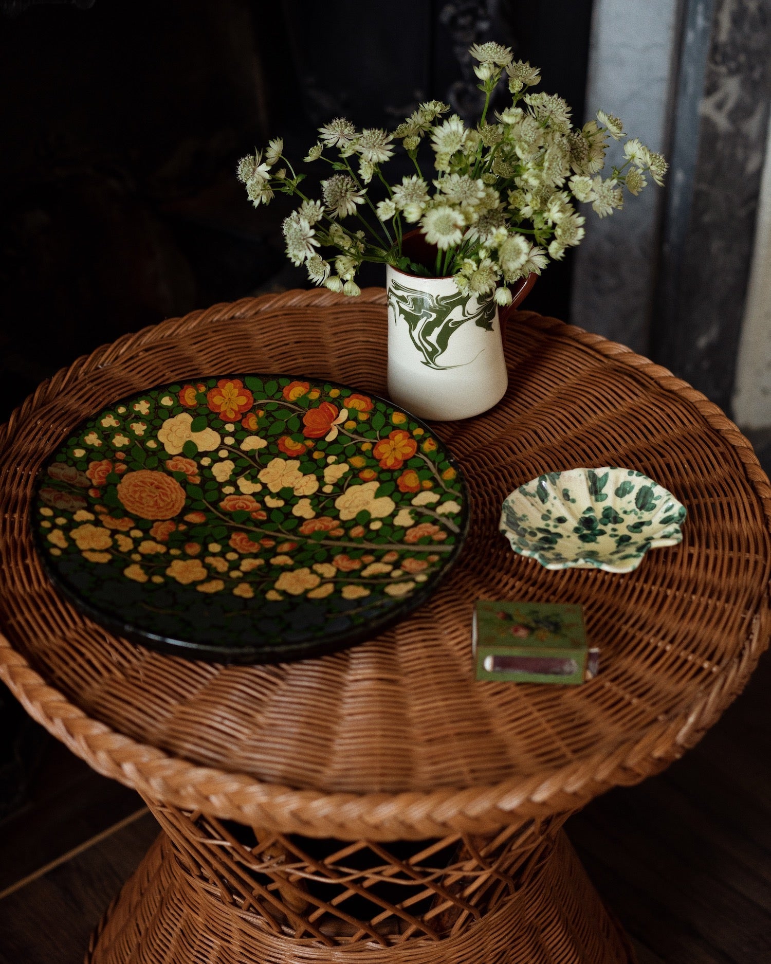A round wicker table displays the Splatter Coquillage Dish Collection by Sharland England, a floral plate, matchbox, and a vase of white flowers—an elegant, handmade-in-Italy scene.