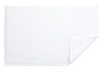 The Reverie Bath Rug by Matouk, a rectangular white cotton rug with a textured surface, is shown flat on a white background with one corner folded up to display its secure non-slip backing.