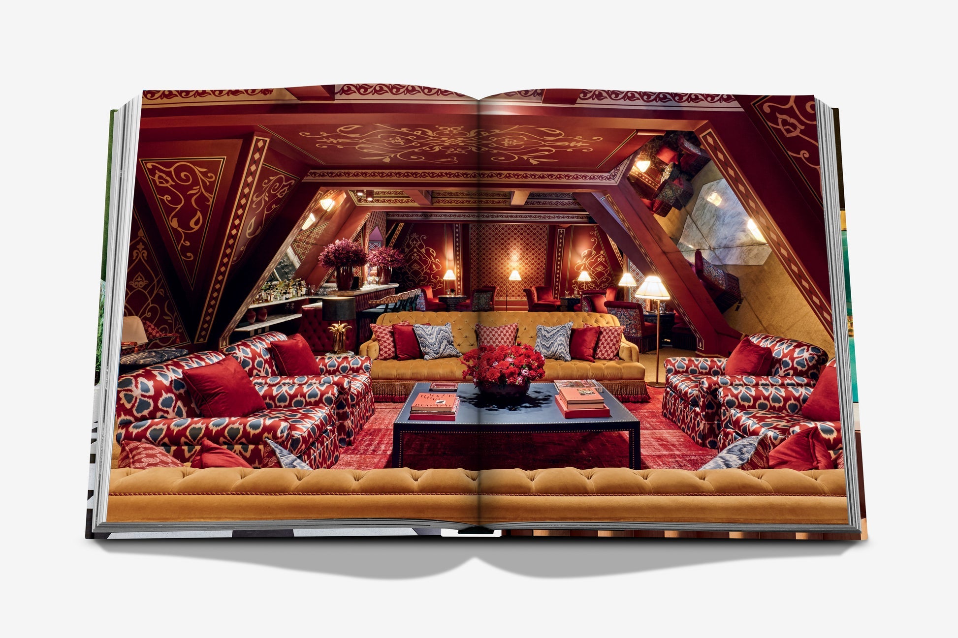 Open book reveals a lavish red-themed living room by Sig Bergamin Eclectic for Assouline, featuring patterned sofas, a coffee table, and decorative cushions.