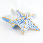 The Herend Starfish Box Collection features a star-shaped ceramic box with a removable lid, adorned with hand-painted blue and gold geometric designs and gold dots on a white background.
