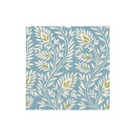 The Samsara Collection by Caspari features a square tile with a blue background and a repeating pattern of white leafy branches and green florals, inspired by eco-friendly napkins and natural designs.