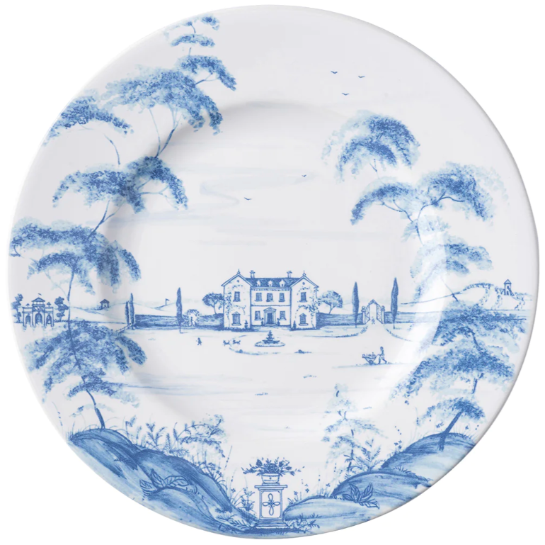 Juliska's Country Estate Delft Blue Dinnerware Collection features a white ceramic plate adorned with a blue pastoral scene of a countryside home, trees, people by a horse stable, and classical architectural elements.
