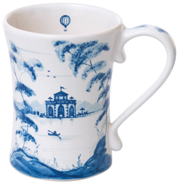 The Juliska Country Estate Delft Blue Dinnerware Collection features a white ceramic mug adorned with a blue pastoral scene—trees, a stable, buildings, a boat on water, and a hot air balloon—perfect for lovers of quiet adventure.