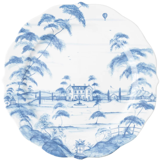 The Juliska Country Estate Delft Blue Dinnerware Collection features a decorative plate adorned with a blue pastoral landscape, large house, horse stable, trees, distant hills, and a hot air balloon—perfect for countryside charm.