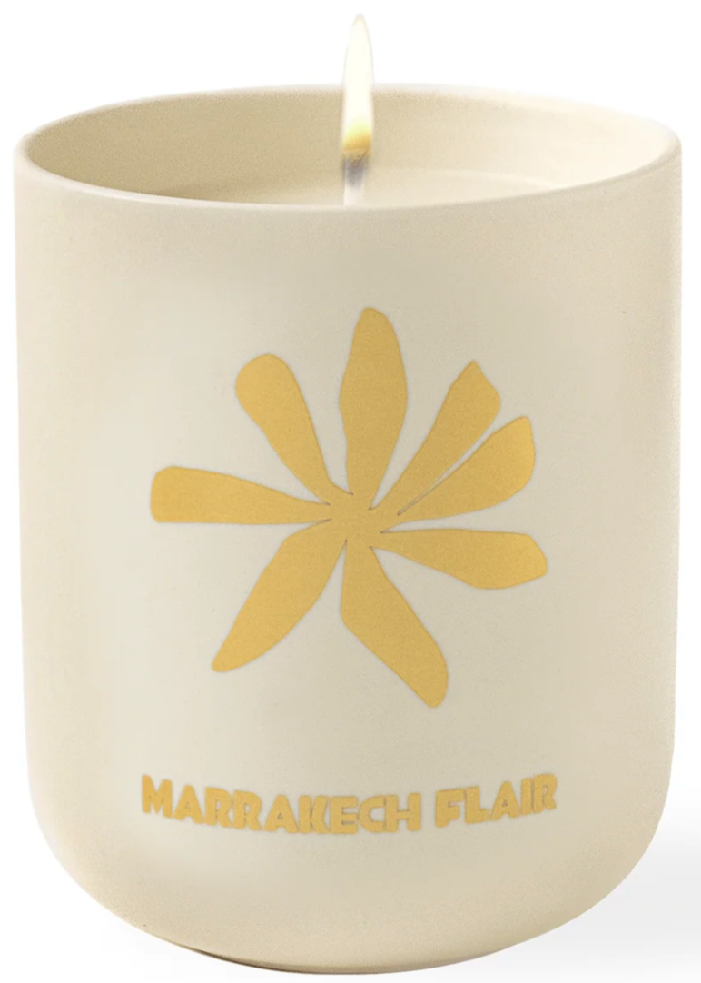 A lit Assouline Travel Candle Marrakech Flair in a cream-colored container with a gold flower design and "marrakech flair" text.