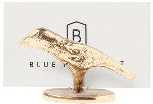 The Hailey Gold Place Card Holder Set by Blue Pheasant features a small, gold-colored bird sculpture with a lustrous finish standing on an oval base, displayed in front of a card with “BLUE B” partially visible.