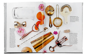 An open book, displaying the "Celebrate Rose" from Graphic Image, elegantly showcases gold and silver bar tools alongside pink flowers and rosé cocktails on a white background, offering ideal party inspiration.