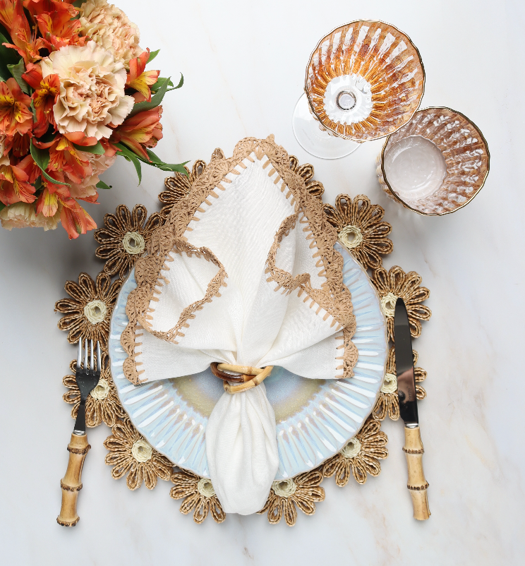 A formal table is set with a Kim Seybert Daisy Placemat, plate, napkin with ring, bamboo-handled fork and knife, two glass goblets, and orange flowers—ideal for elegant outdoor gatherings.