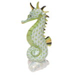 The Herend Sea Horse is a porcelain figurine featuring hand-painted designs, a green scale pattern, gold accents on its tail, fins, and horn, plus a glossy finish. It stands upright on a reflective surface.