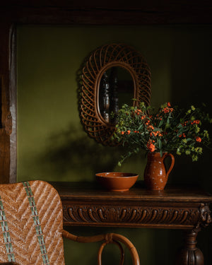 A brown ceramic jug with orange flowers and a matching bowl rest on a carved wooden table beneath the Sharland England Oval Mirror on an olive green wall, with a woven chair in the foreground.