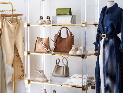 vignette of Hive Boutiques with mannequin wearing blue belted dress over white pants, an etagere with various handbags and shoes, and hanging rack with tan pants