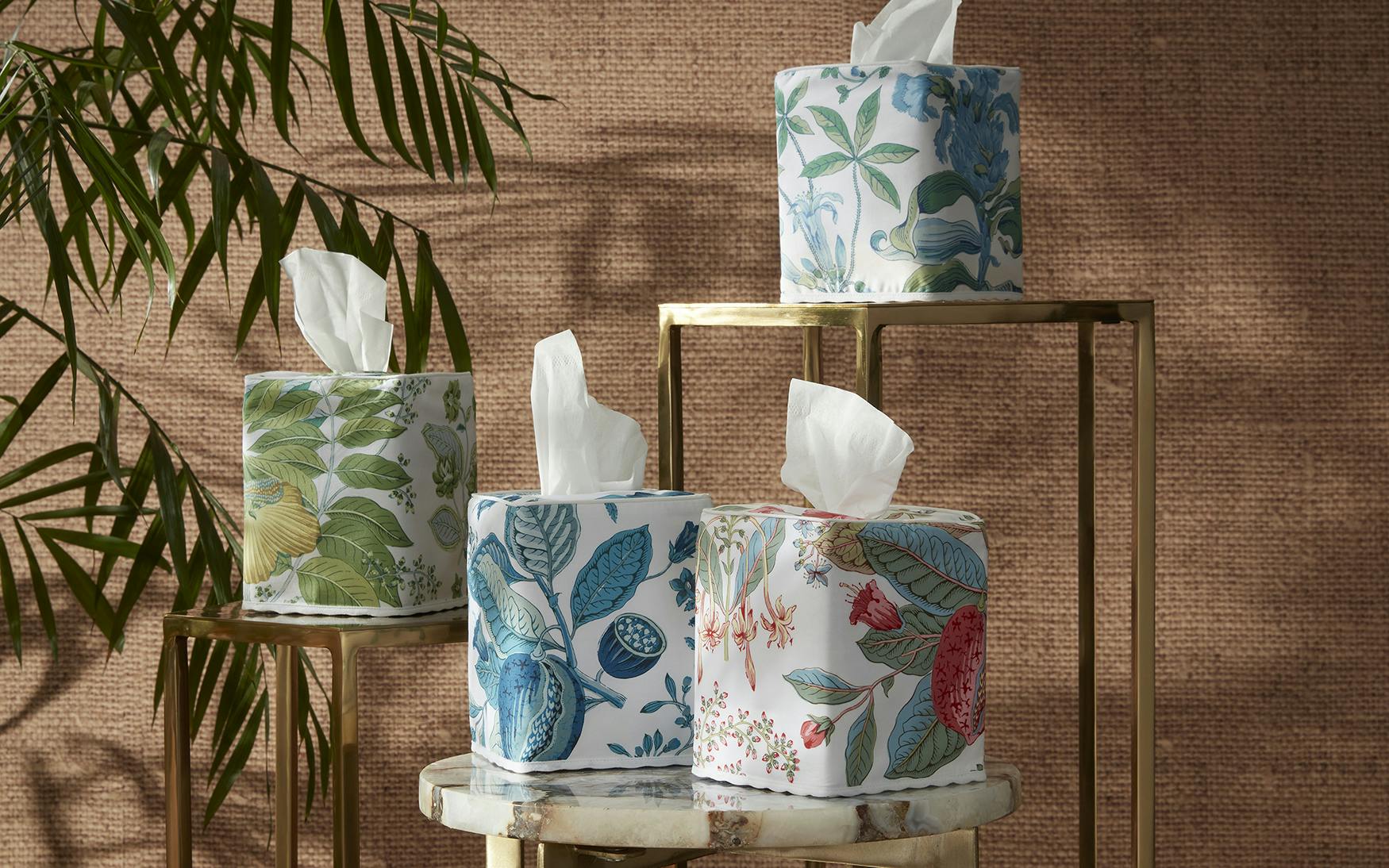 The Matouk Pomegranate Citrus Tissue Box Cover is displayed on stands and a marble table, featuring colorful, pomegranate-inspired botanical designs with a leafy plant in the background.