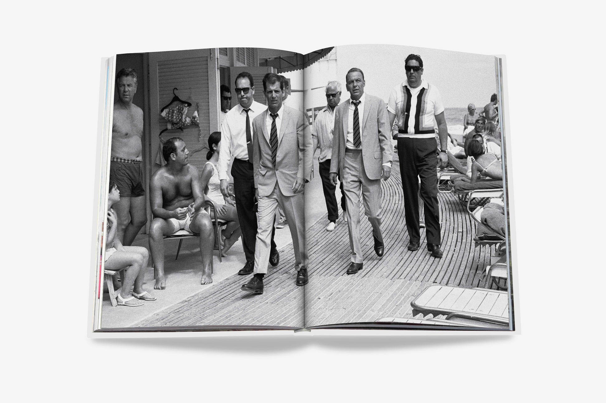 A black and white photo in an open book from Assouline's "The Surf Club" shows men in suits walking on a boardwalk near a beach, surrounded by people in swimwear, capturing the luxury of Miami's iconic club.