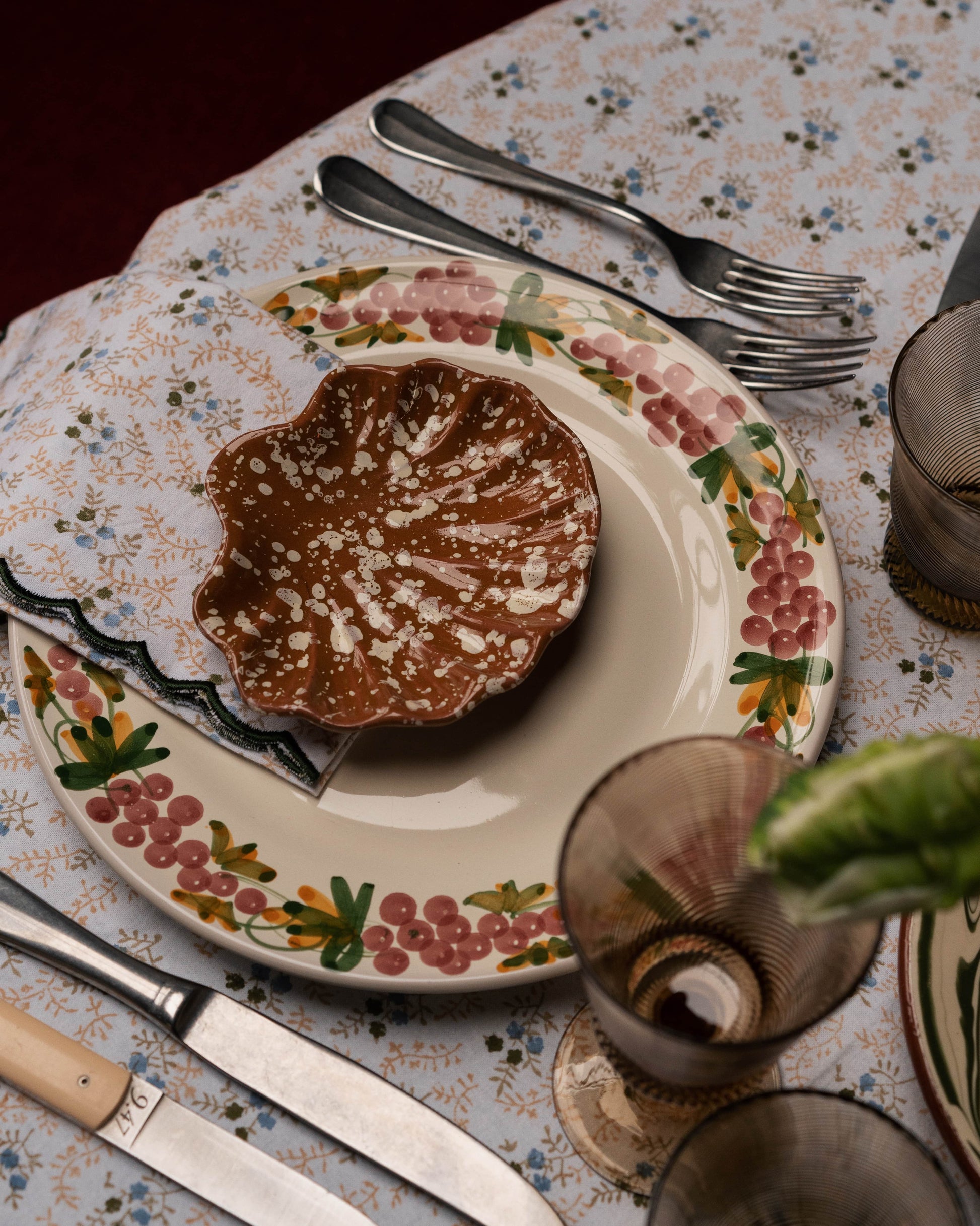 A table setting featuring Sharland England's Splatter Coquillage Dish Collection, silverware, a knife, glasses, and a napkin arranged on a floral tablecloth.
