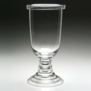 The Tilly Footed Hurricane by William Yeoward Crystal features a clear glass goblet with a thick, round base and cylindrical bowl—ideal for stylish outdoor entertaining or use as a hurricane lamp against a gradient gray background.