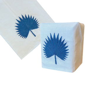 Haute Home’s Palm Leaf Bath Collection, Blue, includes a white Italian linen napkin and hand-embroidered tissue box cover, both adorned with a blue palm leaf motif.