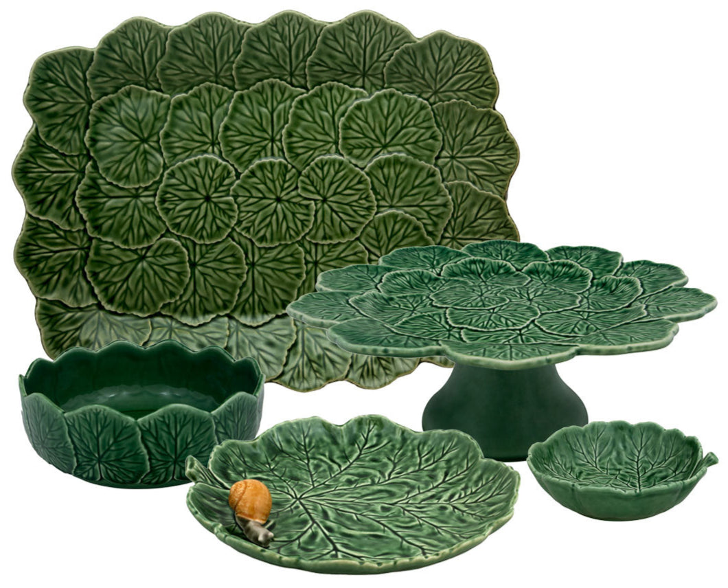 The Hive Geranium Leaf Serveware Collection features green ceramic dishes and trays with leaf patterns—a platter, bowl, cake stand, and plate are shown together. A snail is resting on one of the plates.