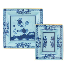 Two square ceramic trays from the Oriente Italiano Iris Tray Collection by Ginori 1735, showcasing light blue backgrounds, dark blue carnation floral motifs, yellow rims, and geometric border designs.