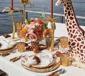 A table set for four features Kim Seybert's Royal Palm Placemats, gold accents, a floral centerpiece, pinecone napkin holders with hand-applied beads, and a large giraffe figurine by the water.