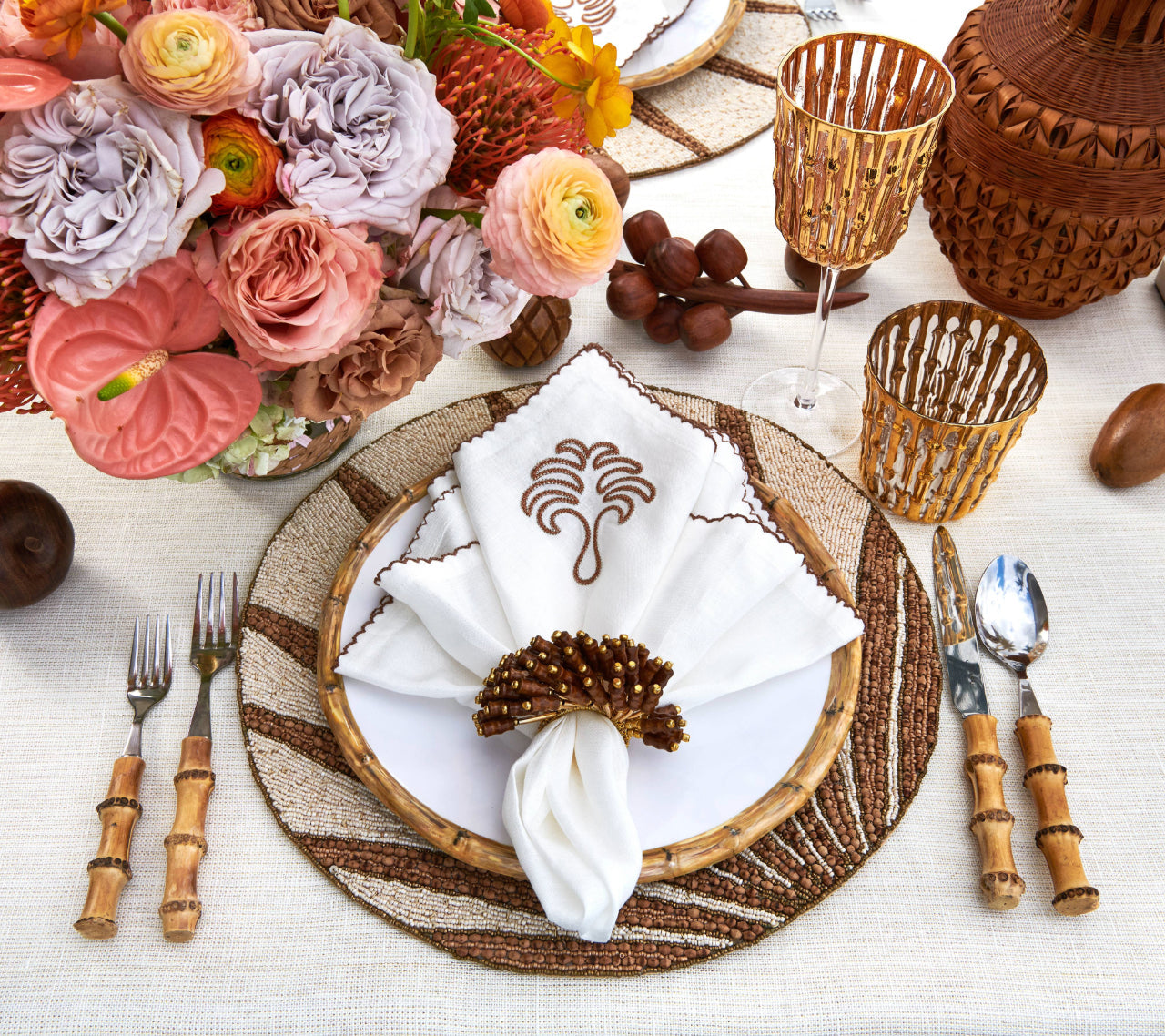 A table setting features a white plate, patterned placemat, bamboo-handled cutlery, embroidered napkin with the Kim Seybert Royal Palm Napkin Ring, floral centerpiece, and amber glassware for chic tropical style.
