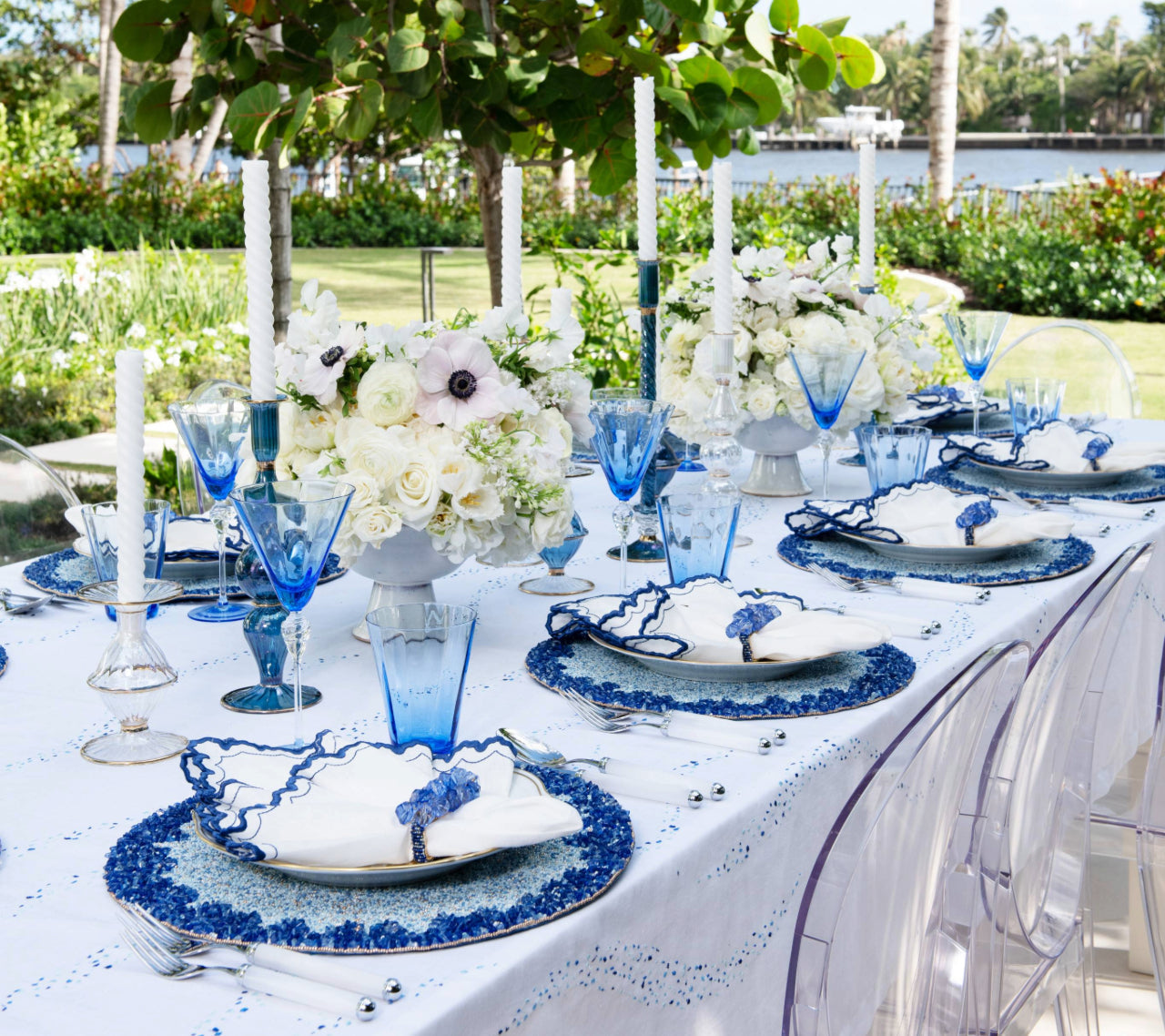 An outdoor dining table features blue and white plates, cobalt blue beads, blue glassware, white flowers, tall white candles, clear chairs, and the Kim Seybert Lagoon Placemat for an ocean-inspired touch amid lush greenery and water views.