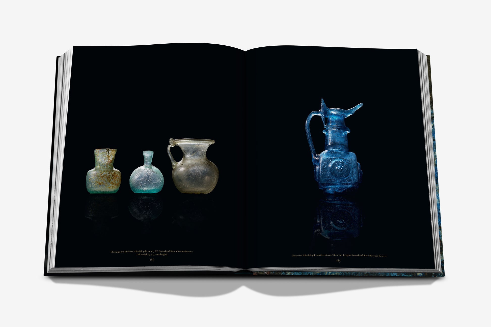 An open book from Assouline, titled *Uzbekistan: Masterpieces of the Silk Road*, showcases images of four ancient glass vessels against a black background – three adorn the left page, while an intricately designed blue pitcher from Uzbekistan graces the right page, echoing treasures of the Silk Road.