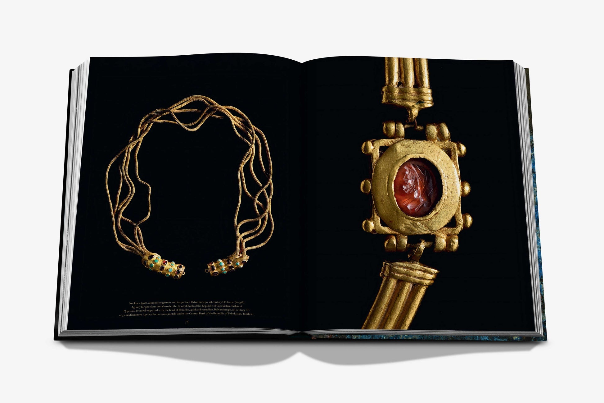 An Assouline book, "Uzbekistan: Masterpieces of the Silk Road," is open, presenting images of gold jewelry reminiscent of historical treasures: a multi-strand necklace on the left and an archaeological artifact with a red gemstone set in gold on the right, celebrating Uzbekistan's rich heritage.