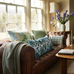 A brown leather sofa with Varun Euro Decorative Pillows by John Robshaw, solid cushions, a gray throw blanket, and a wooden coffee table with books sits by large windows and a vase of purple flowers.
