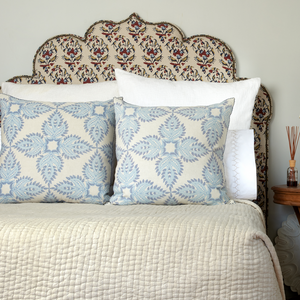 A bed with a hand block printed fabric headboard, two white pillows, two Verdin Lapis Euro Pillows by John Robshaw in blue and white, a cream quilt, and a small wooden side table topped with a reed diffuser.