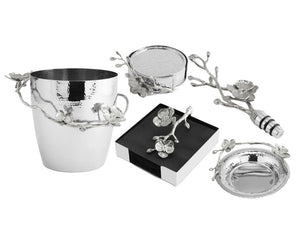 The White Orchid Barware Collection by Michael Aram features silver items with floral and branch designs inspired by orchids, including an ice bucket, coaster, bottle stopper, and napkin holder that reflect purity and grace.