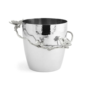 The Michael Aram White Orchid Barware Collection features a hammered silver ice bucket with a decorative handle adorned with elegant floral and branch details inspired by orchids.