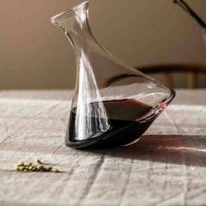 The Simon Pearce Walden Rolling Wine Decanter, partially filled with red wine, rests on a light tablecloth—ideal for aerating your favorite wines.