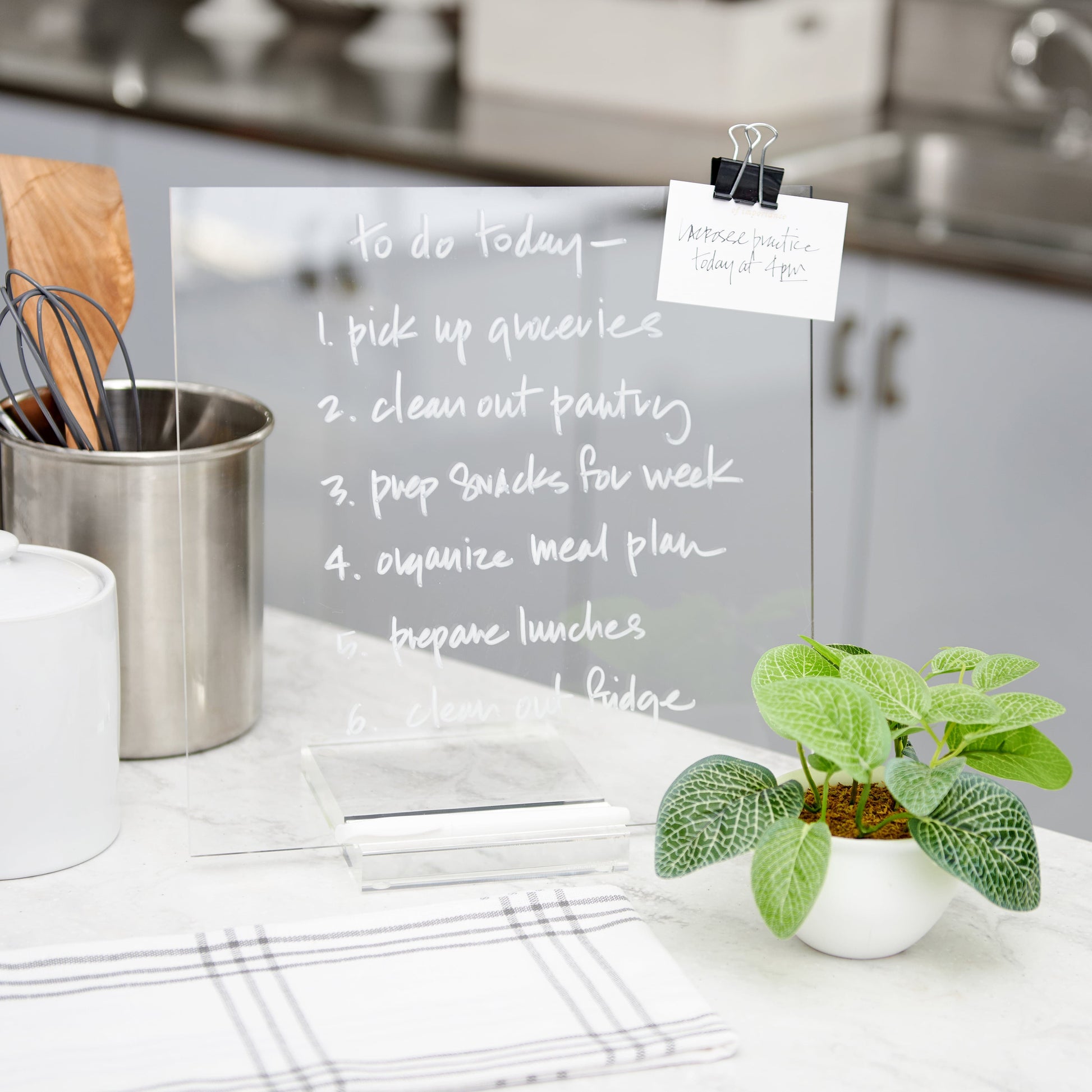 A Russell & Hazel Acrylic Memo Tablet sits on the kitchen counter with a vibrant handwritten to-do list from a wet erase marker. Nearby, a lush plant thrives, and an elegant note is clipped above, exemplifying modern design.