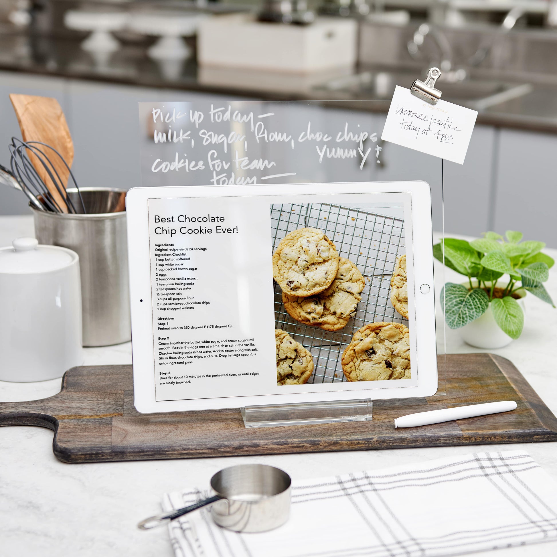 The Russell & Hazel Acrylic Memo Tablet with its modern design stands on the kitchen counter displaying a chocolate chip cookie recipe, while baking ingredients and utensils wait nearby, along with a wet erase marker for quick notes.