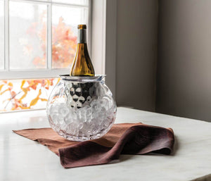 The Simon Pearce Hampton Chiller, crafted with handmade glassblowing techniques, holds a wine bottle and ice cubes on a white table by the window, set atop a brown cloth.