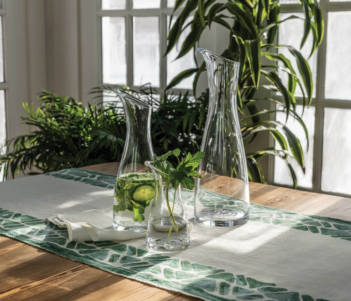 Three Simon Pearce Barre Carafe Collection pieces, one filled with water, cucumber, and mint, rest on a wooden table with a leafy runner in a bright room featuring greenery and large windows.