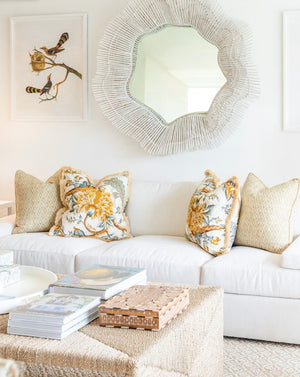 A white sofa with down-filled yellow floral cushions stands in a bright room. A circular woven mirror and bird artwork hang on the wall. A wicker ottoman holds books and a decorative box, adding charm to this inviting space enhanced by the Le Grand Corail Bleu Pillow with Fringe by Associated Design, custom handmade in the USA.